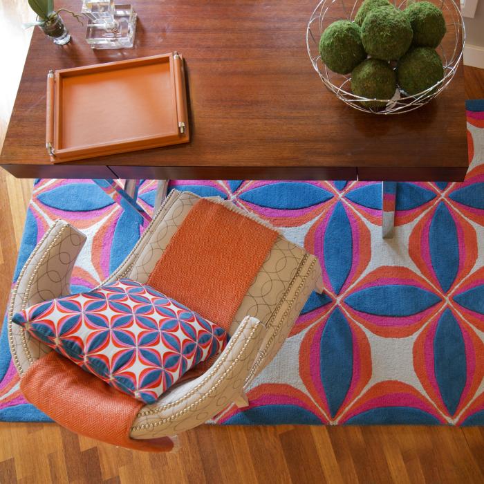 J Company room with rug and pillow in shades of blue, orange and white, cream chair with orange throw, wooden desk and décor