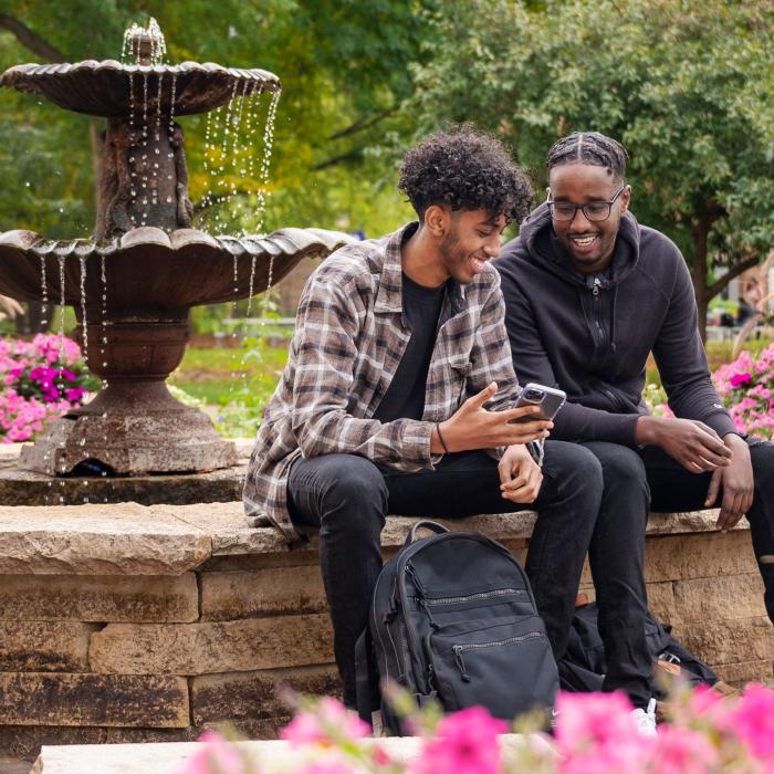 Two male Winona State University students enjoying a campus courtyard with fountain and flowers in bloom